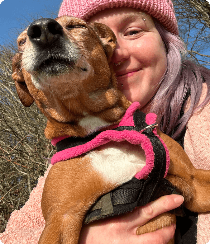 Hannah in a pink bobble hat being licked on the cheek by a happy tan and white dog